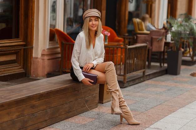 Guapa modelo rubia sexy con blusa blanca y skrt de cuero y botas altas hasta el muslo de bloque de punto en beige