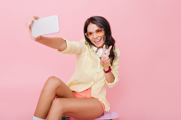 Guapa mujer europea con bronceado bronceado haciendo selfie en smartphone. Alegre niña adorable en chaqueta amarilla tomando una foto de sí misma con el signo de la paz.