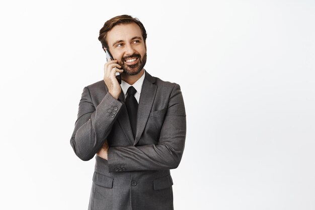 Guapo agente inmobiliario sonriendo hablando por teléfono móvil y mirando el banner de espacio vacío en la esquina superior derecha de pie sobre fondo blanco.