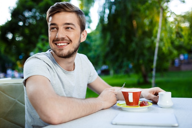 Guapo alegre sonriente joven sentado en la mesa de café al aire libre con una taza de café.