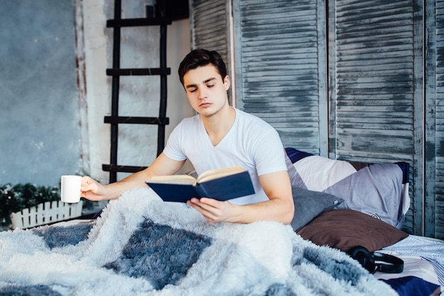 Guapo, apuesto joven tendido sin camisa en su cama, sosteniendo una taza de café o té mientras lee un libro