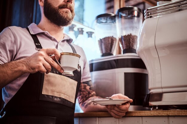 Guapo barista con delantal bebiendo café durante el almuerzo apoyado en un mostrador en la cafetería