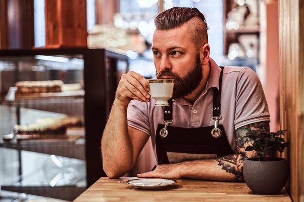 Guapo barista en delantal bebiendo café durante el almuerzo sentado en una mesa en la cafetería