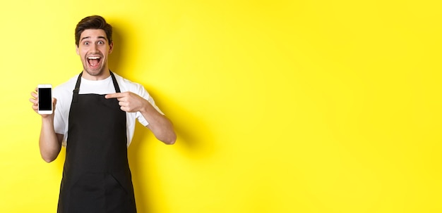 Foto gratuita guapo barista en delantal negro apuntando con el dedo a la pantalla móvil que muestra la aplicación y sonriendo de pie ov