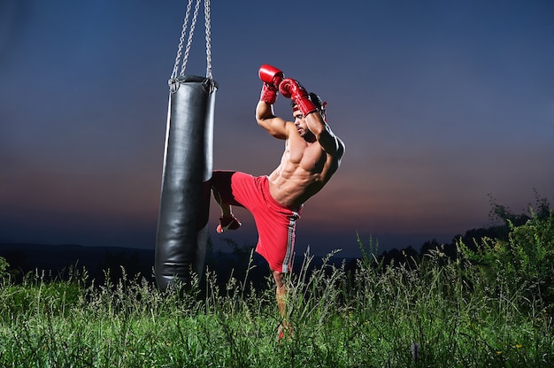 Guapo boxeador joven musculoso sin camisa que trabaja con un saco de boxeo al aire libre copyspace hermosa puesta de sol en el fondo naturaleza estilo de vida deportes atleta activo entrenamiento de masculinidad atlética.