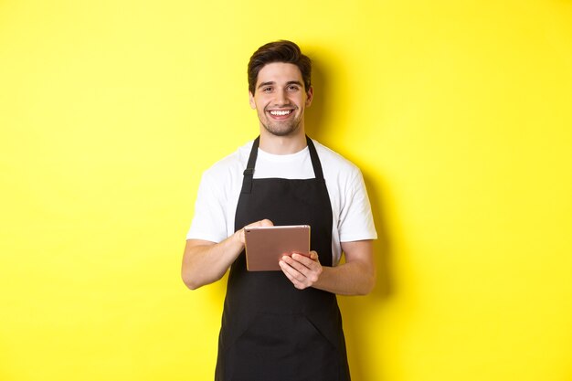 Guapo camarero tomando órdenes, sosteniendo tableta digital y sonriendo, vestido con uniforme de delantal negro, de pie sobre fondo amarillo.