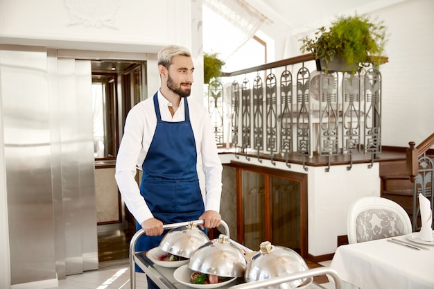 El guapo camarero vestido con el uniforme sirve platos calientes en el restaurante.