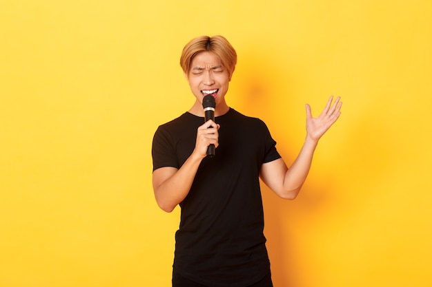Guapo cantante masculino asiático, chico coreano cantando canciones en el karaoke en el micrófono con pasión, de pie sobre la pared amarilla