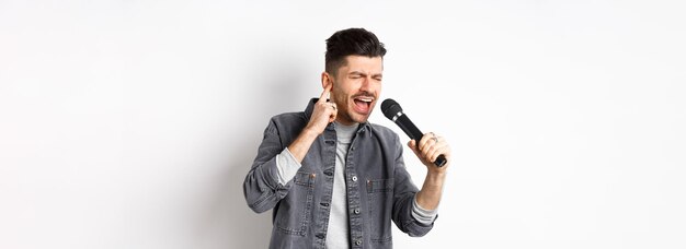 Guapo cantante tratando de alcanzar una nota alta cantando en el micrófono y cerrar una oreja de pie en blanco b