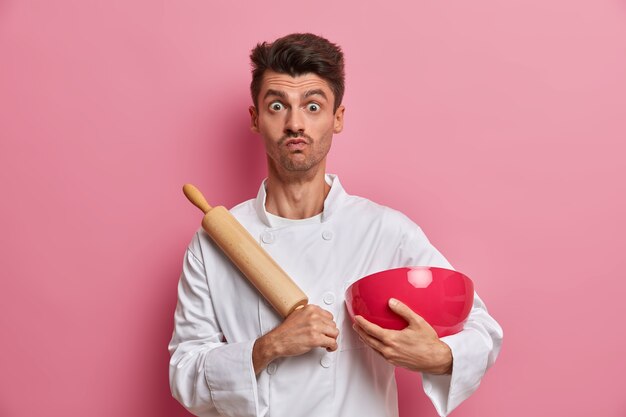 Guapo chef masculino con expresión de sorpresa, cocina comida en la cocina, sostiene un rodillo y un tazón, prepara masa fresca, viste uniforme blanco