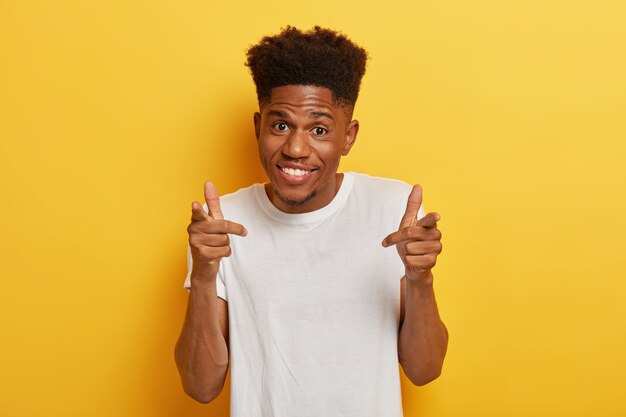 Guapo chico afroamericano rizado señala con el dedo índice con alegría, sonríe agradablemente, está de buen humor, te recoge en su alegre compañía, viste camiseta blanca, modelos sobre pared amarilla.