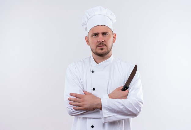 Guapo cocinero joven en serio en uniforme de chef de pie con postura cerrada y sosteniendo un cuchillo aislado en el espacio en blanco