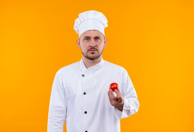 Guapo cocinero joven en serio en uniforme de chef sosteniendo tomate y mirando aislado en el espacio naranja