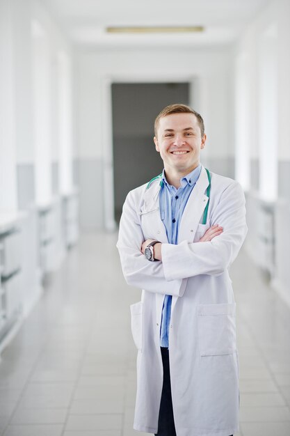 Guapo y confiado joven médico en bata blanca con estetoscopio posando en el hospital