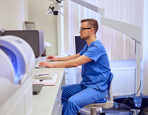 Guapo dentis masculino usando computadora en una habitación con equipo médico en el fondo.