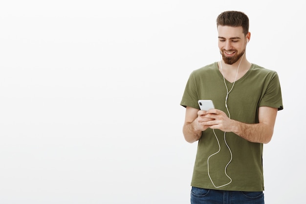 Guapo, elegante, barbudo, con audífonos, sosteniendo un teléfono inteligente y sonriendo alegremente en la pantalla del móvil, viendo videos en un lugar público, usando audífonos, no molestar a la gente