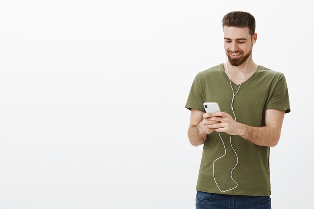 guapo, elegante, barbudo, con audífonos, sosteniendo un teléfono inteligente y sonriendo alegremente en la pantalla del móvil, viendo videos en un lugar público, usando audífonos, no molestar a la gente