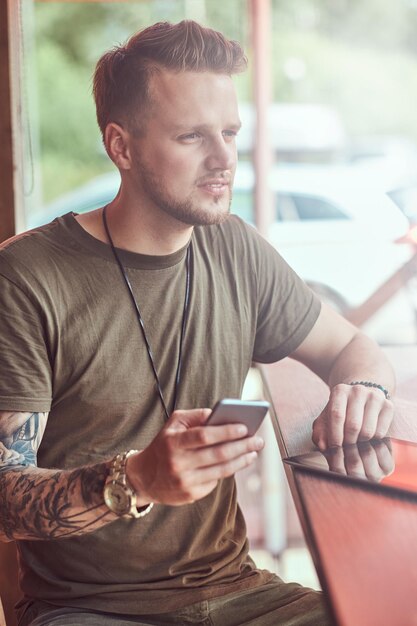 Guapo y elegante hipster se sienta en una mesa en un café al borde de la carretera, sostiene el teléfono inteligente.