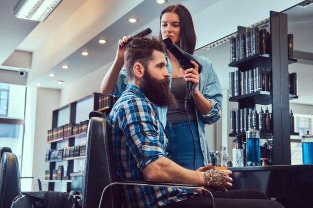 Guapo y elegante hombre barbudo con un tatuaje en el brazo vestido con una camisa de franela sosteniendo jugo mientras la peluquera usa un secador de pelo en una barbería.