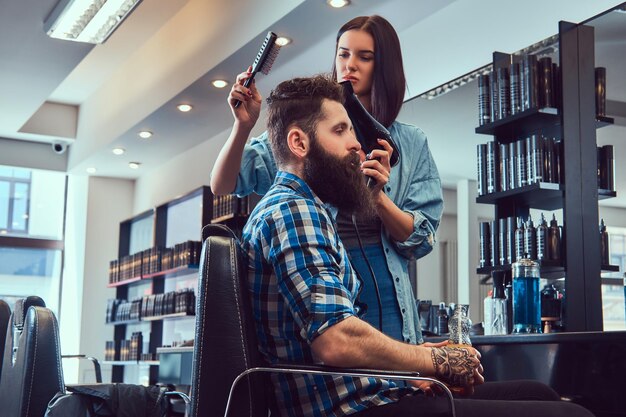 Guapo y elegante hombre barbudo con un tatuaje en el brazo vestido con una camisa de franela sosteniendo jugo mientras la peluquera usa un secador de pelo en una barbería.