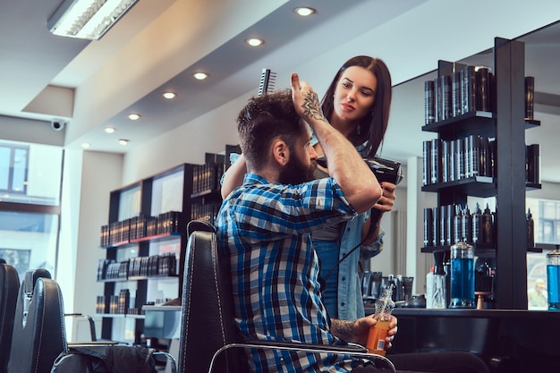 Guapo y elegante hombre barbudo con un tatuaje en el brazo vestido con una camisa de franela sosteniendo jugo mientras la peluquera usa un secador de pelo en una barbería.