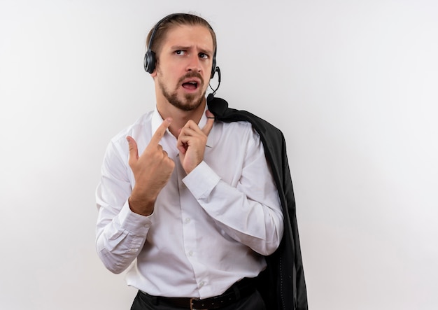 Guapo empresario sosteniendo la chaqueta sobre el hombro con auriculares con un micrófono mirando a un lado confundido de pie sobre fondo blanco.