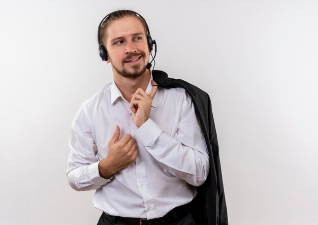 Guapo empresario sosteniendo la chaqueta sobre el hombro con auriculares con un micrófono mirando a un lado con una sonrisa de confianza de pie sobre fondo blanco.