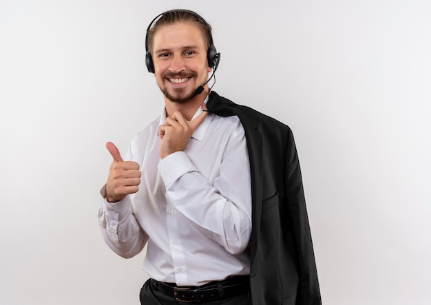 Foto gratuita guapo empresario sosteniendo la chaqueta sobre el hombro con auriculares con un micrófono sonriendo mostrando los pulgares para arriba sobre fondo blanco.