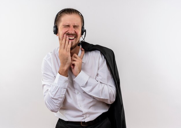 Guapo empresario sosteniendo la chaqueta sobre el hombro con auriculares con un micrófono tocando la mejilla con dolor de muelas de pie sobre fondo blanco.