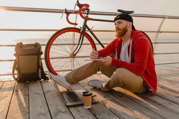 Guapo estilo hipster hombre barbudo que trabaja como autónomo en línea en una computadora portátil con mochila y bicicleta en el amanecer de la mañana junto al mar mochilero de viajero de estilo de vida activo y saludable