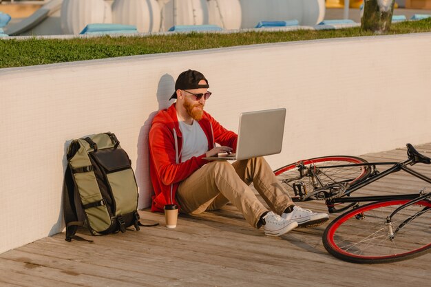 Guapo estilo hipster hombre barbudo que trabaja como autónomo en línea en una computadora portátil con mochila y bicicleta en el amanecer de la mañana junto al mar mochilero de viajero de estilo de vida activo y saludable