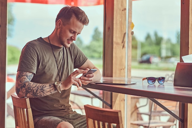 Guapo hipster con estilo se sienta en una mesa en un café al borde de la carretera, leyendo el mensaje en el teléfono inteligente.