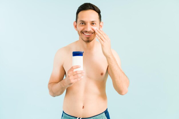 Guapo hispano de unos 20 años no quiere quemarse con el sol en la playa. Hombre latino en traje de baño aplicando loción de protección solar en la cara