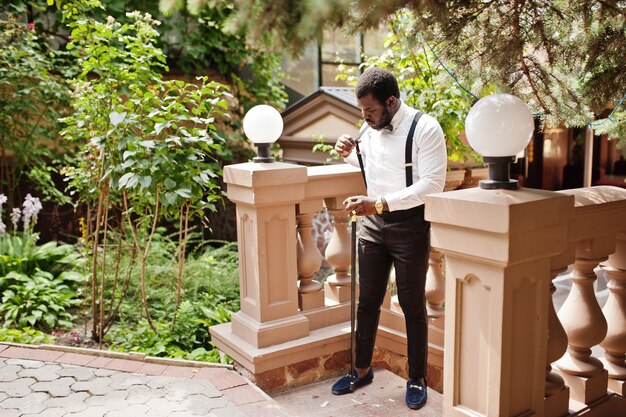 Guapo hombre afroamericano de moda en traje formal pajarita y tirantes bastón