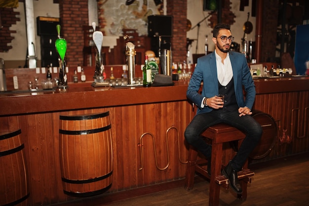 Foto gratuita guapo hombre árabe bien vestido con vaso de whisky y cigarro posado en el pub