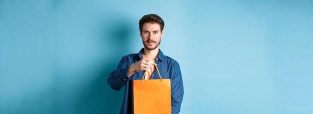 Foto gratuita guapo hombre barbudo estirar la mano con una bolsa de compras naranja haciendo un regalo de pie sobre backgr azul