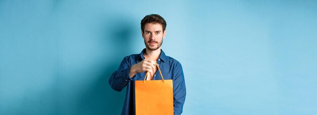 Guapo hombre barbudo estirar la mano con una bolsa de compras naranja haciendo un regalo de pie sobre backgr azul