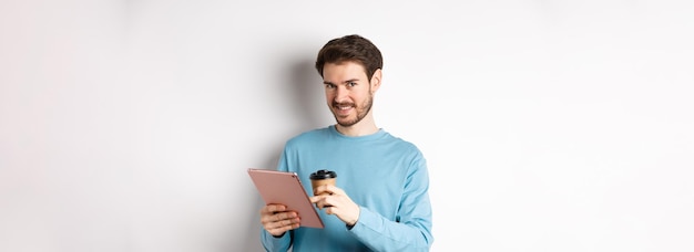 Foto gratuita guapo hombre barbudo sonriendo a la cámara bebiendo café y leyendo en tableta digital de pie sobre w