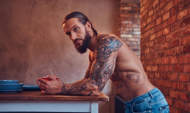 Guapo hombre barbudo tatuado con un elegante corte de pelo y cuerpo musculoso, bebe café, apoyado en una mesa.