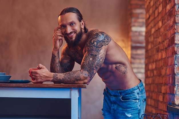 Guapo hombre barbudo tatuado con un elegante corte de pelo y cuerpo musculoso, bebe café, apoyado en una mesa.