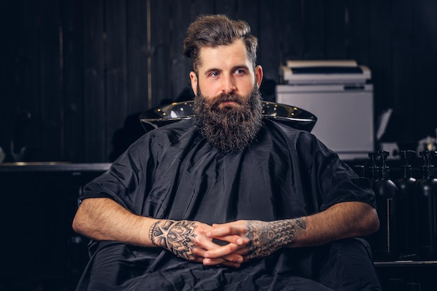 Guapo hombre barbudo con un tatuaje en los brazos antes de lavarse el cabello en un salón de peluquería.