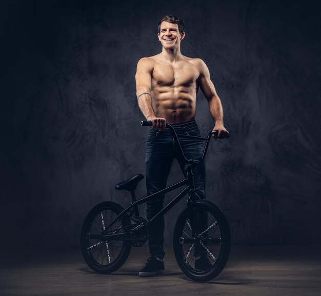 Guapo hombre sin camisa sonriente en camiseta y jeans de pie con BMX. Aislado en un fondo oscuro.