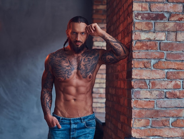 Foto gratuita guapo hombre sin camisa tatuado con un elegante corte de pelo y barba, de pie apoyado contra una pared de ladrillos en una cocina.