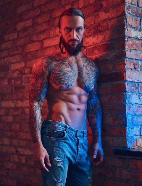 Foto gratuita guapo hombre sin camisa tatuado con un elegante corte de pelo y barba, de pie contra una pared de ladrillos en una habitación con un interior de loft.