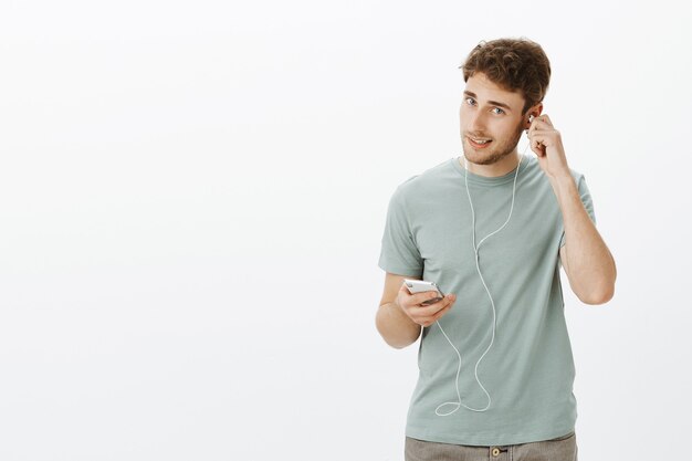 Guapo hombre caucásico confiado con cerdas, poniéndose los auriculares y sosteniendo el teléfono inteligente, escogiendo una canción y preparándose para caminar con música en los oídos