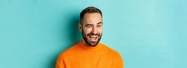 Guapo hombre confiado guiñando un ojo a la cámara sonriendo descarado de pie en suéter naranja contra backg azul