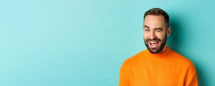 Foto gratuito guapo hombre confiado guiñando un ojo a la cámara sonriendo descarado de pie en suéter naranja contra backg azul