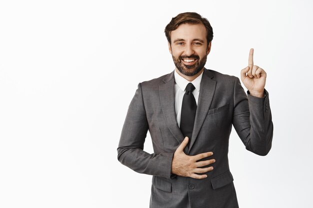Guapo hombre corporativo en traje apuntando con el dedo hacia arriba sosteniendo la mano en su vientre y sonriendo satisfecho concepto de salud y gente de negocios fondo blanco.