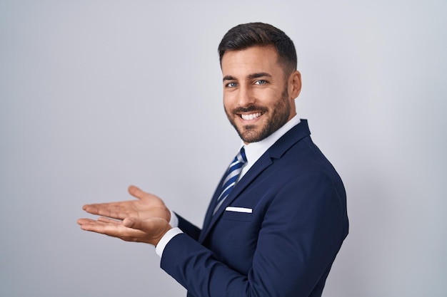 Guapo hombre hispano vestido con traje y corbata apuntando a un lado con las manos abiertas mostrando el espacio de la copia, presentando un anuncio sonriendo emocionado feliz