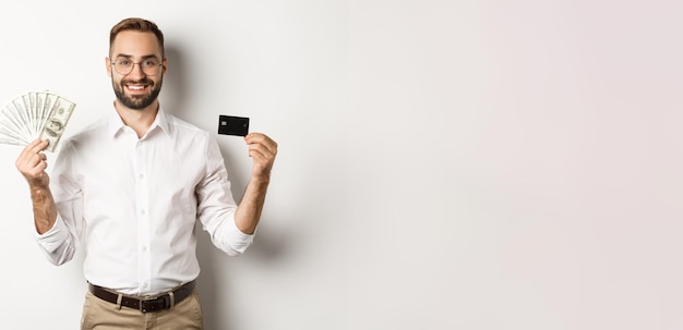 Foto gratuita guapo hombre de negocios mostrando tarjeta de crédito y dinero dólares sonriendo satisfecho de pie sobre blanco ba